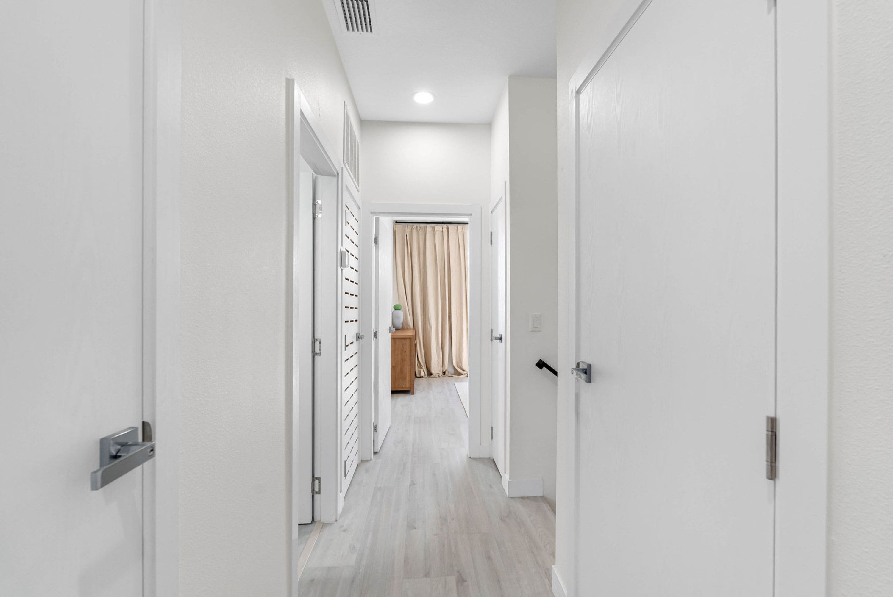 A hallway with white walls and doors, leading to a room with a wooden floor and a curtain.