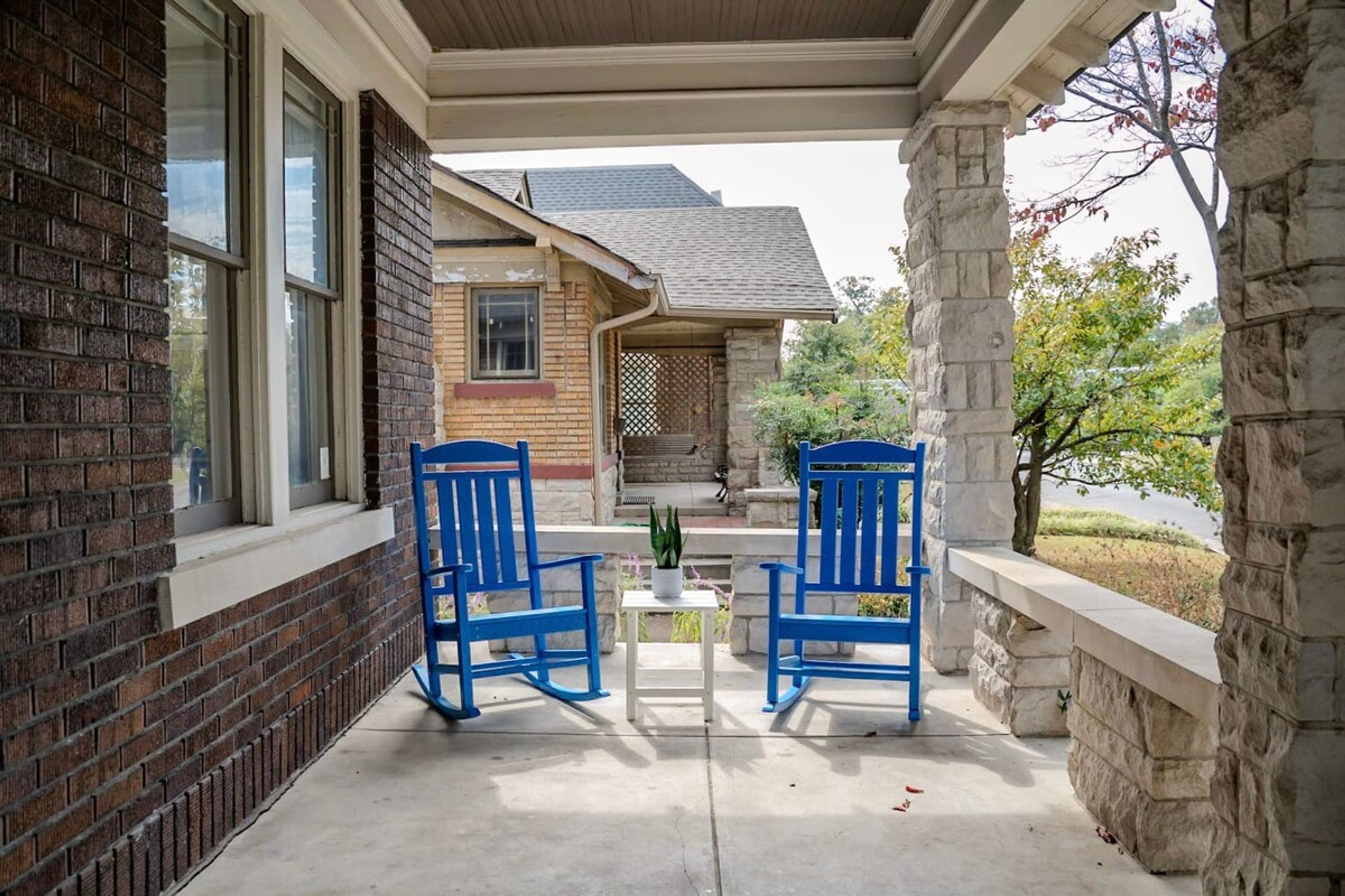 A covered porch with two blue chairs and a small table, a brick wall, and a stone pillar.