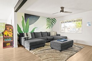 Un salon moderne avec un canapé d’angle, un petit pouf, un tapis à poils longs et une fresque murale représentant de grandes feuilles vertes.