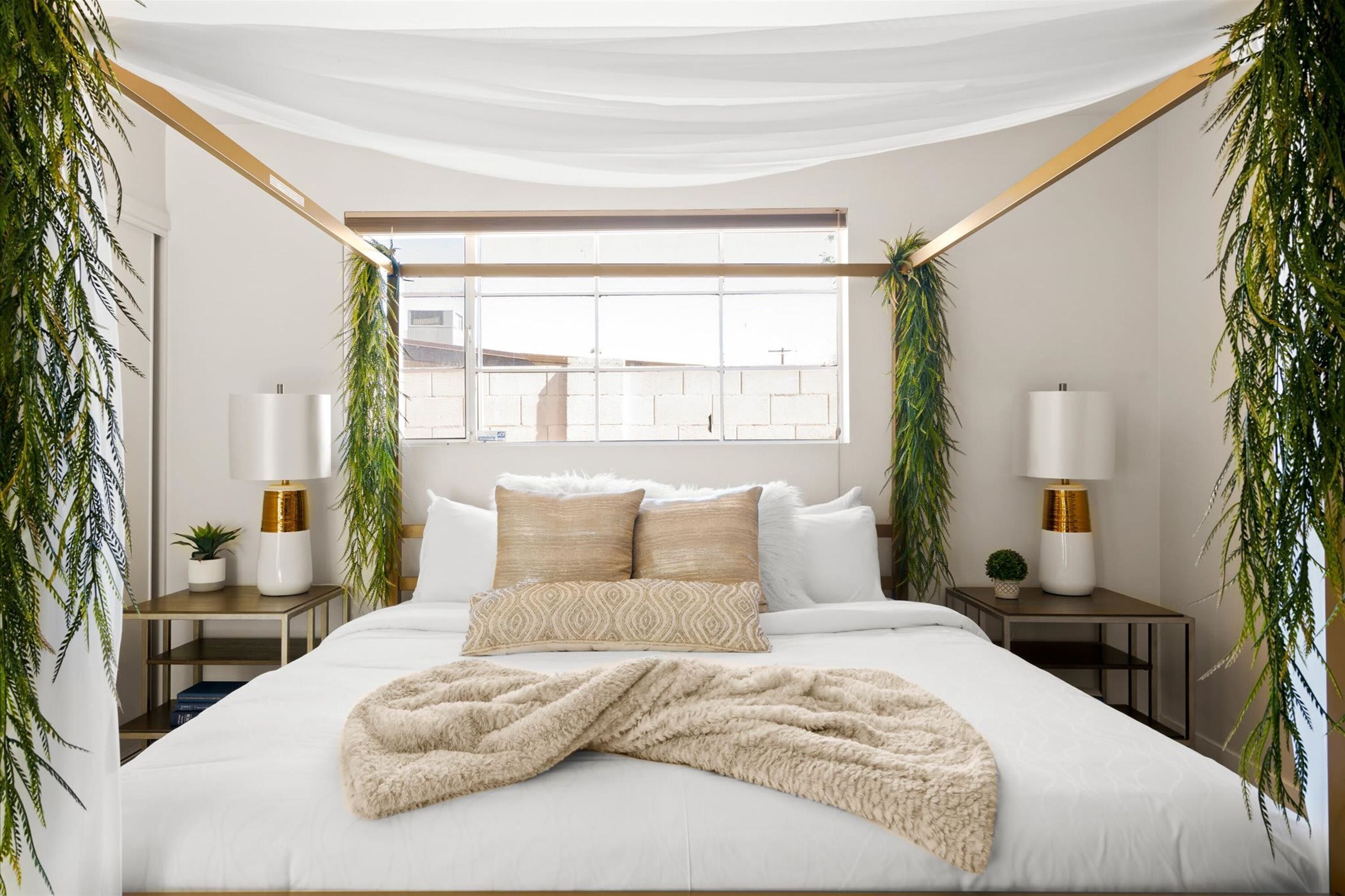 Une chambre avec un grand lit, deux tables de chevet, une fenêtre et des plantes suspendues.