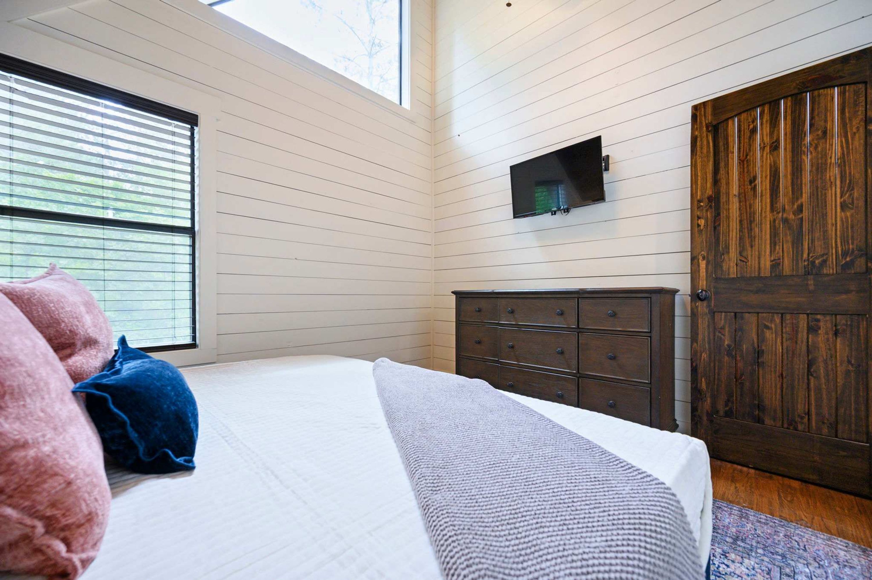 A bedroom with a bed, a dresser, a wooden door, and a window with blinds.