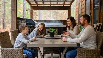 A family of four seated at a round table with a potted plant, in a covered outdoor area with a grill in the background.