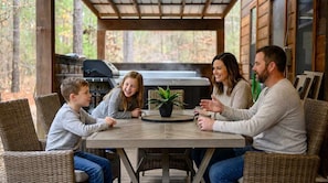 Una familia de cuatro sentada en una mesa redonda con una planta en maceta, en un espacio al aire libre techado con una parrilla al fondo.