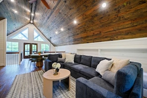 Un loft spacieux avec un plafond haut, un canapé d’angle gris foncé, une table basse ronde et une vue sur l’extérieur grâce à de grandes fenêtres.
