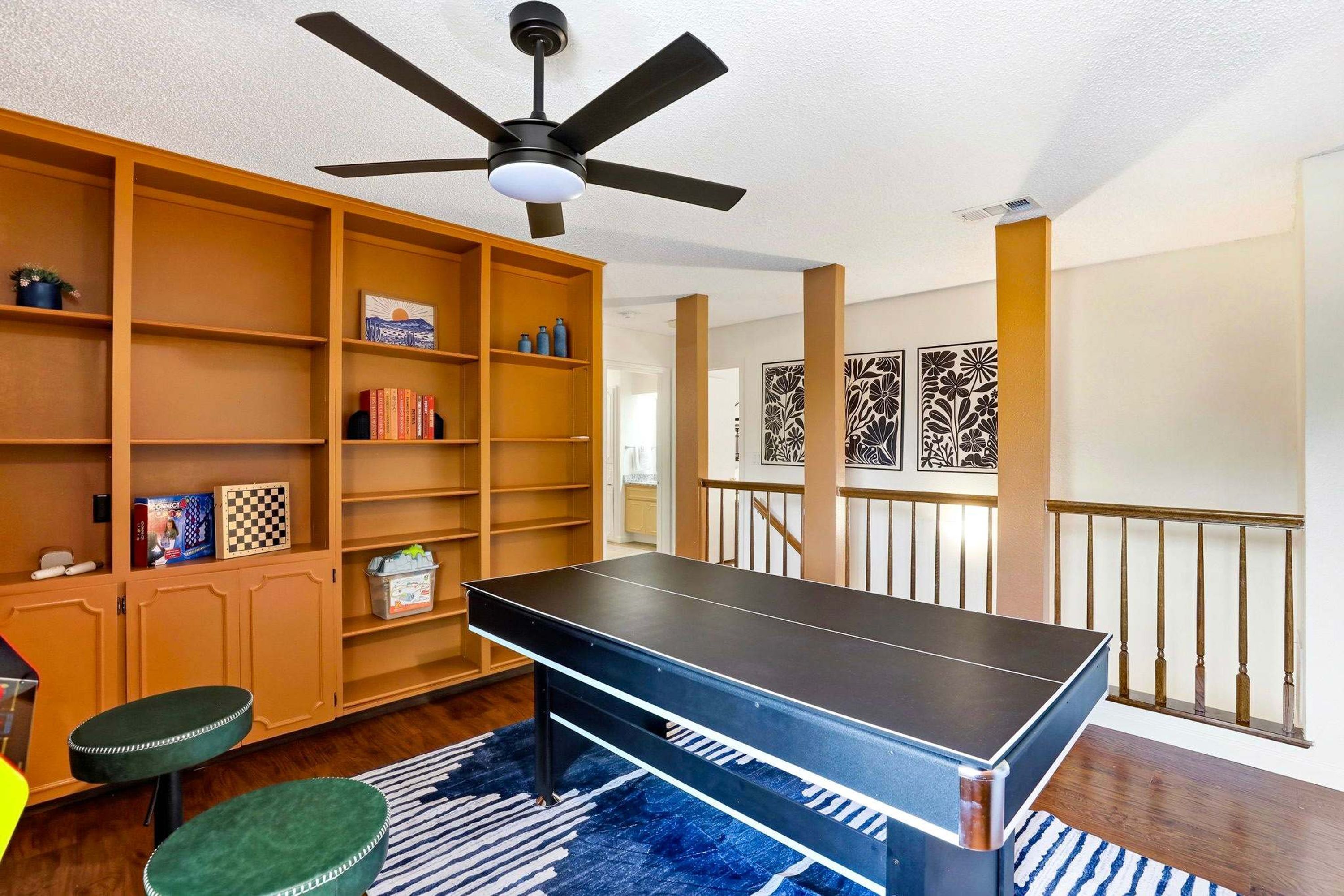 A room with a ping pong table, wooden bookshelves, a ceiling fan, and a staircase with decorative panels.