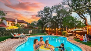 A swimming pool with a curved edge, surrounded by a wooden deck and a hedge. - Elegant Escape Sleeps 14 Games Pool Fire Pit (San Antonio)