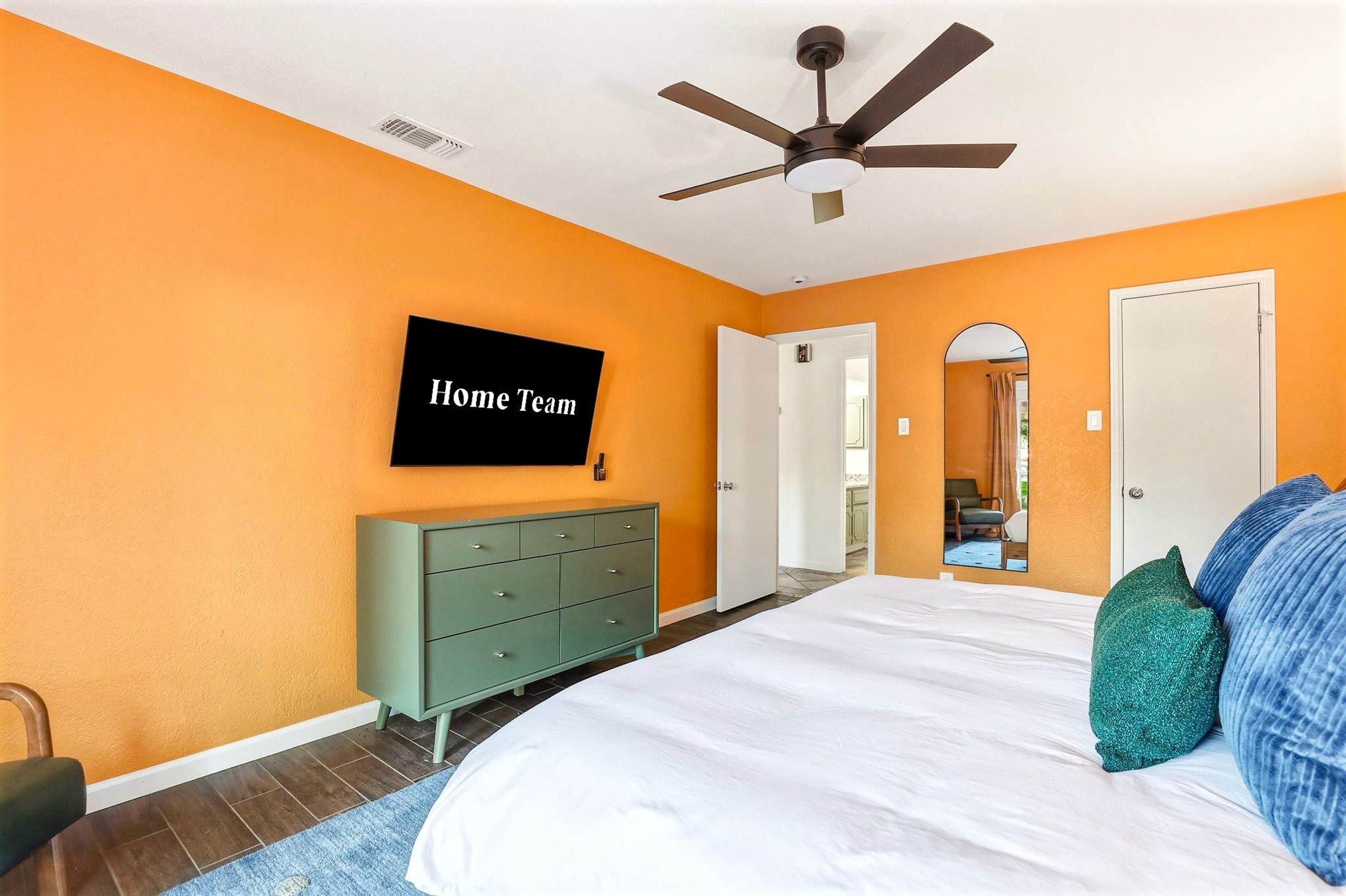 Ein Schlafzimmer mit oranger Wand, einem Bett mit weißer Bettwäsche, einer grünen Kommode, einem Deckenventilator und einem an der Wand montierten Fernseher, der 'Home Team' anzeigt.