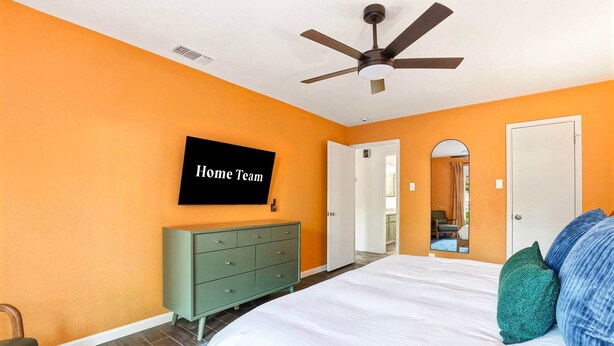 A bedroom with an orange wall, a bed with white bedding, a green dresser, a ceiling fan, and a TV mounted on the wall displaying 'Home Team'. - Elegant Escape Sleeps 14 Games Pool Fire Pit (San Antonio)