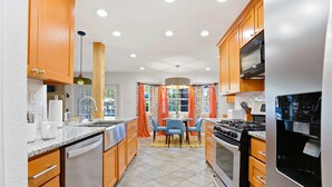 A modern kitchen with wooden cabinets, a stainless steel refrigerator, and a dining area with orange curtains. - Elegant Escape Sleeps 14 Games Pool Fire Pit (San Antonio)