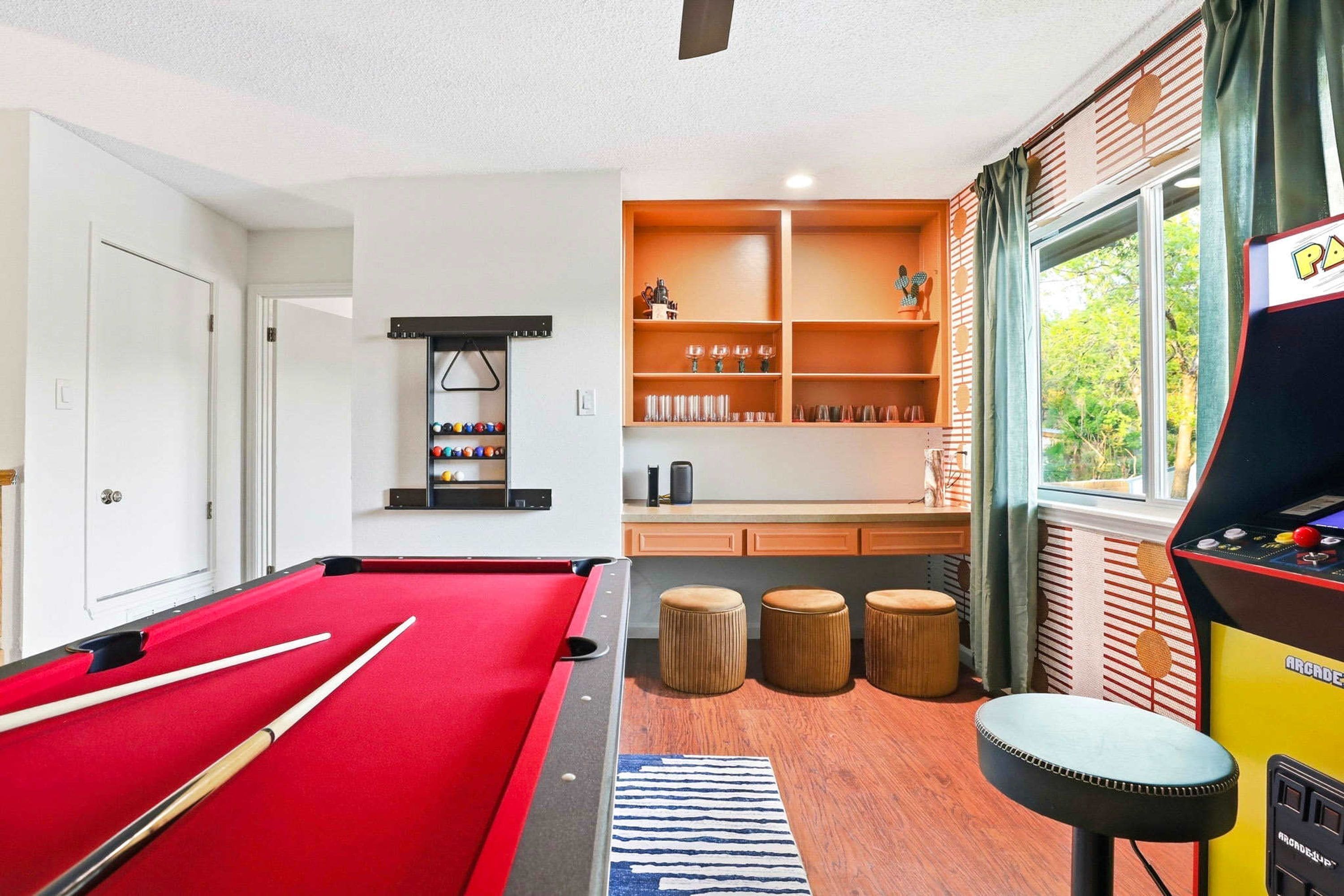 A pool table with cues, a billiards rack, and stools in a room with a window, shelves with glasses, and a pool table.