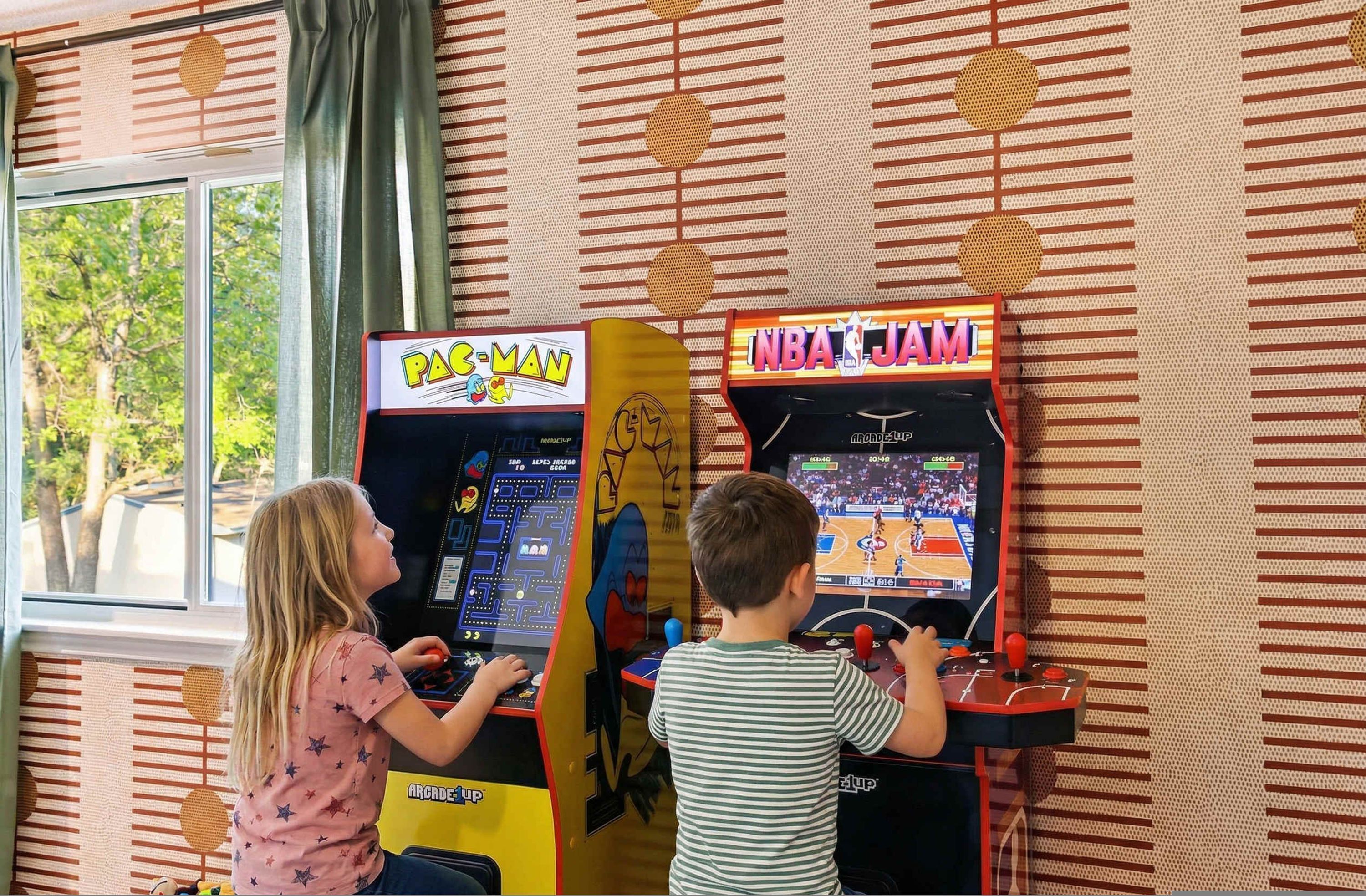 Two arcade gaming machines, one labeled 'Pac-Man' and the other 'NBA Jam,' in a room with patterned wallpaper and a window.