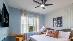 A bedroom with a bed, a desk, a chair, a ceiling fan, a window with curtains, and a wall-mounted picture. - Austin Compound Pickleball Pool Sauna (Austin)