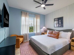 A bedroom with a bed, a desk, a chair, a ceiling fan, a window with curtains, and a wall-mounted picture. - Austin Compound Pickleball Pool Sauna (Austin)