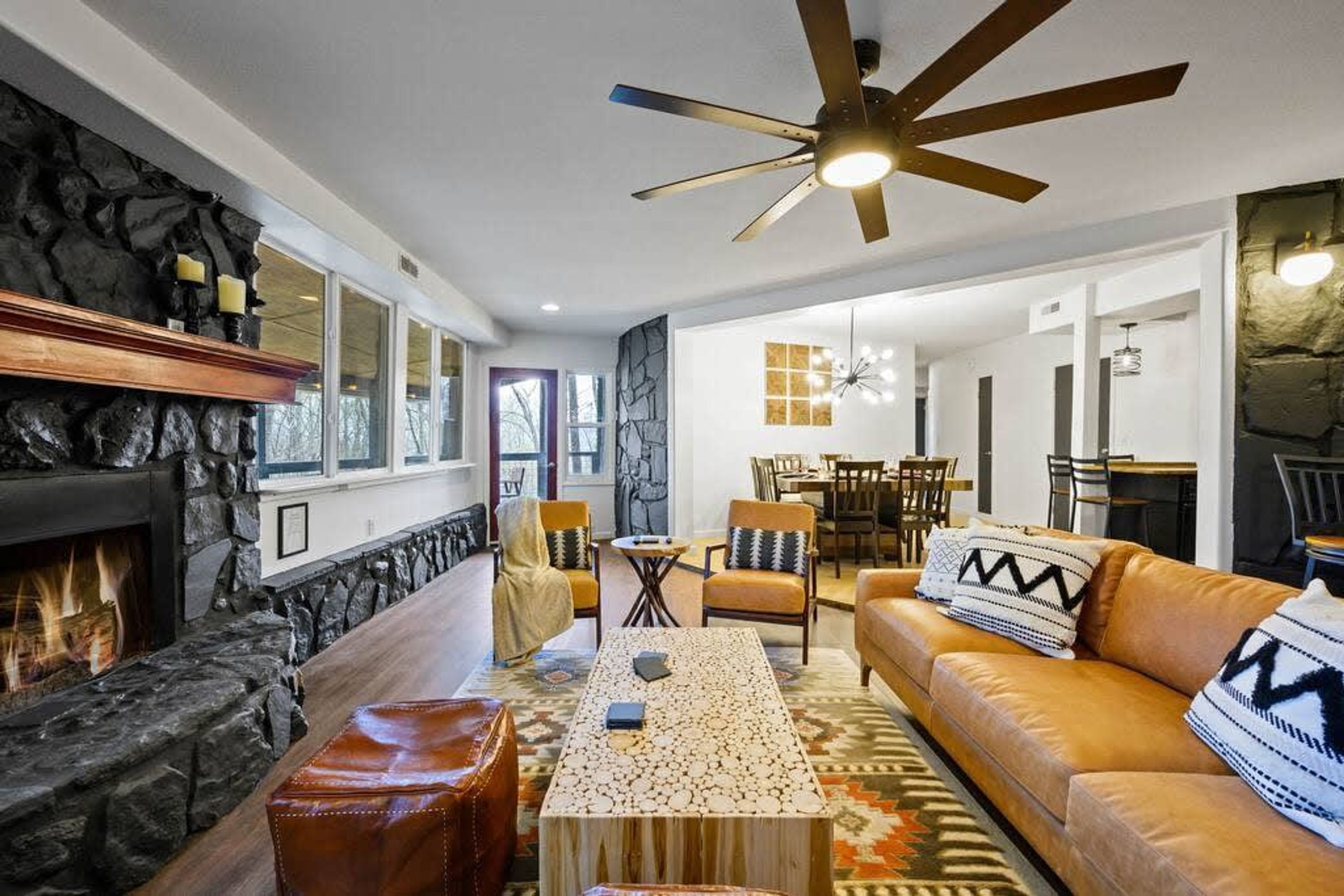Un salon moderne doté d’une cheminée, d’un mur d’accent en pierre, d’un pouf en cuir, d’une table basse à motifs et d’un ventilateur de plafond.