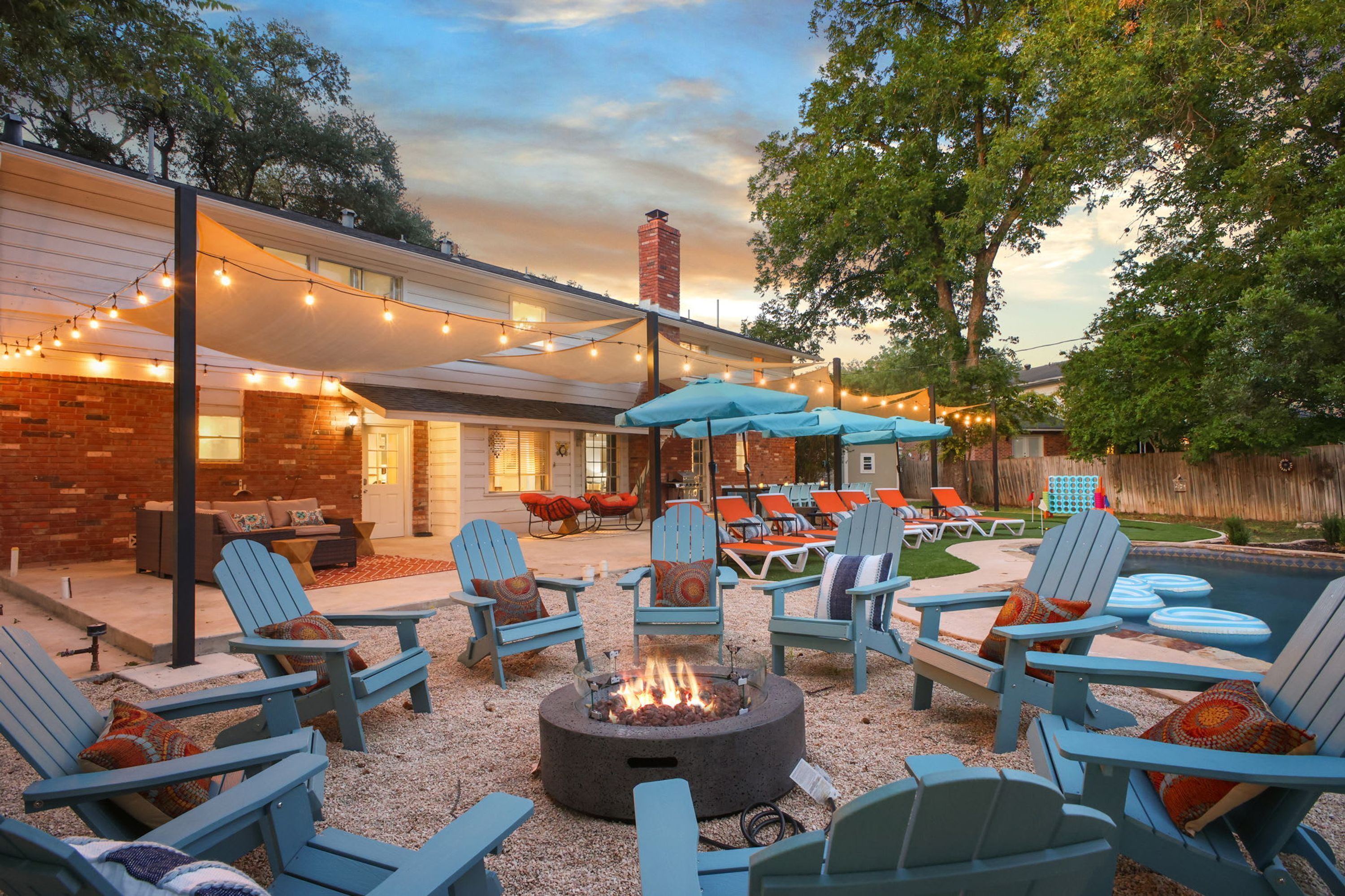 An outdoor patio area with Adirondack chairs, a fire pit, string lights, and a pool.
