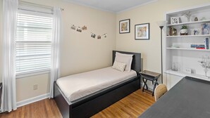 Un dormitorio con cama, mesita de noche, lámpara, estante con libros y adornos, y ventana con persianas.