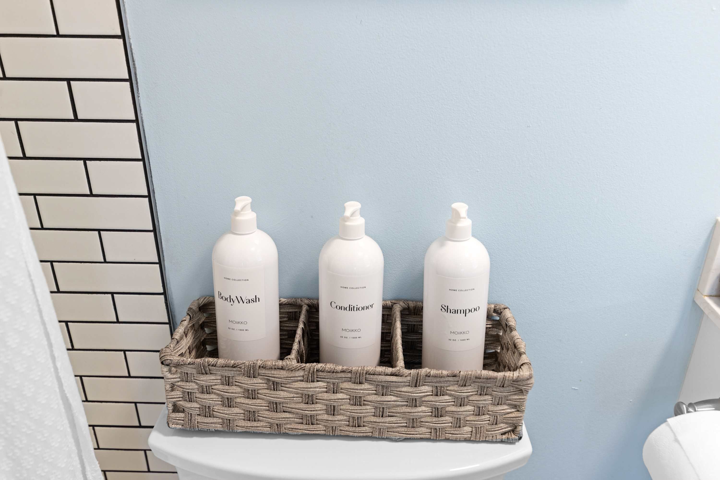 A bathroom with a white toilet, a white wall, and a woven basket containing three toiletry bottles labeled 'BodyWash,' 'Conditioner,' and 'Shampoo.'