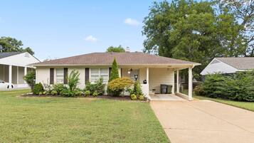 A single-story house with a covered porch, a front yard with a lawn and shrubs, and a neighboring house.