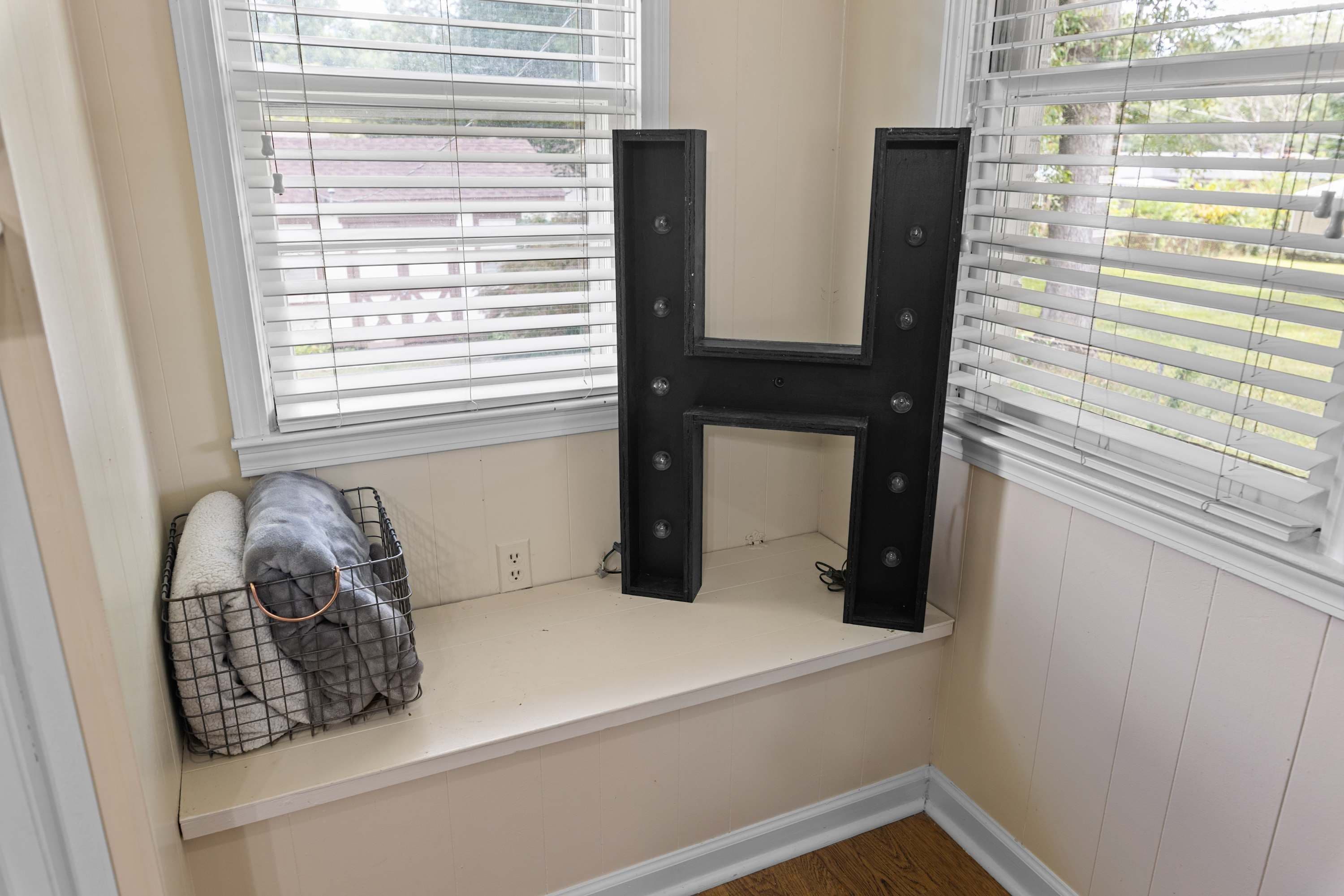 A window seat with a black letter H decoration, a wire basket with a gray blanket, and a white wall.