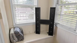 Asiento junto a la ventana con una decoración en forma de letra H negra, una cesta de alambre con una manta gris y una pared blanca.