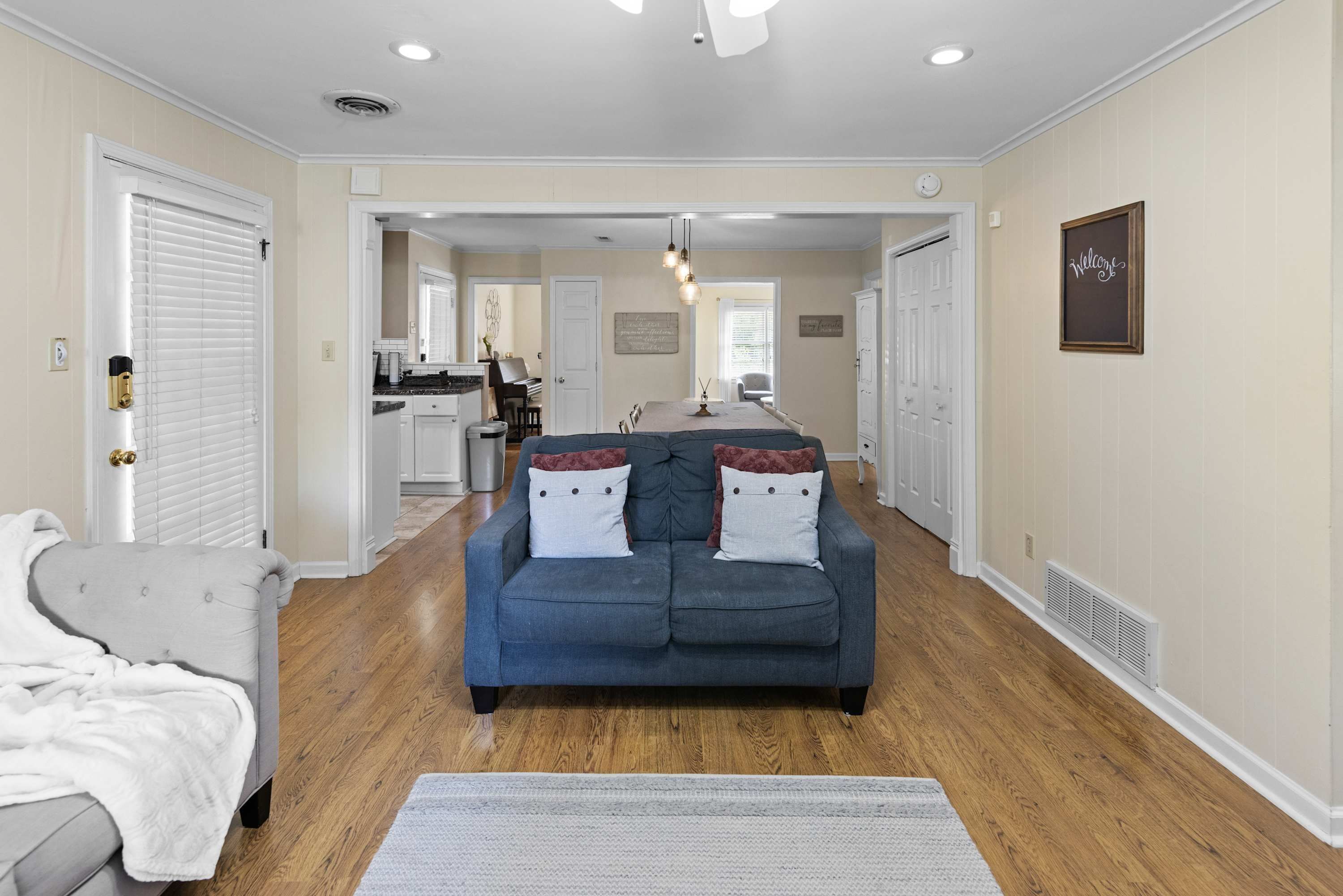 A spacious living room with a blue sofa, a white armchair, and a grey couch. There is a kitchen in the background with a dining area.