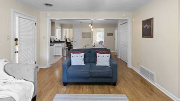 A spacious living room with a blue sofa, a white armchair, and a grey couch. There is a kitchen in the background with a dining area.