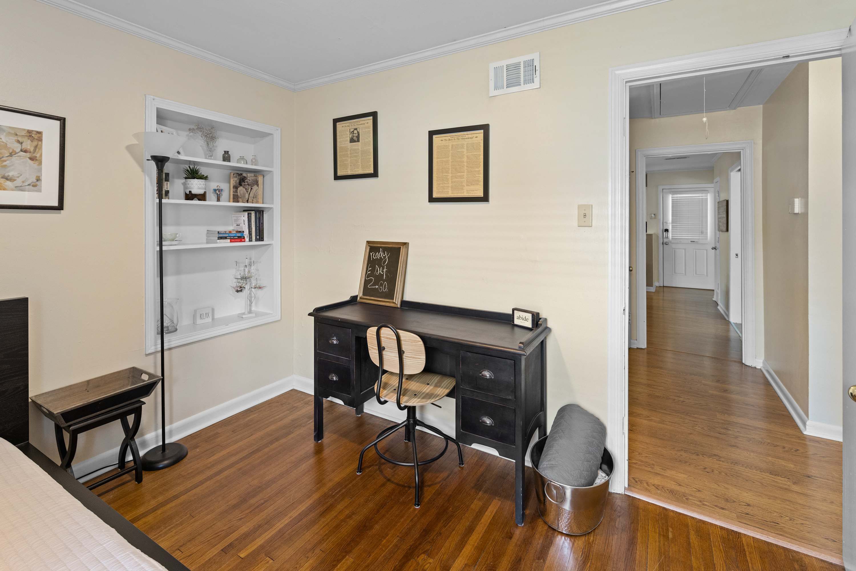 A room with a wooden floor, a desk with a chair, a bookshelf, and a framed picture on the wall.