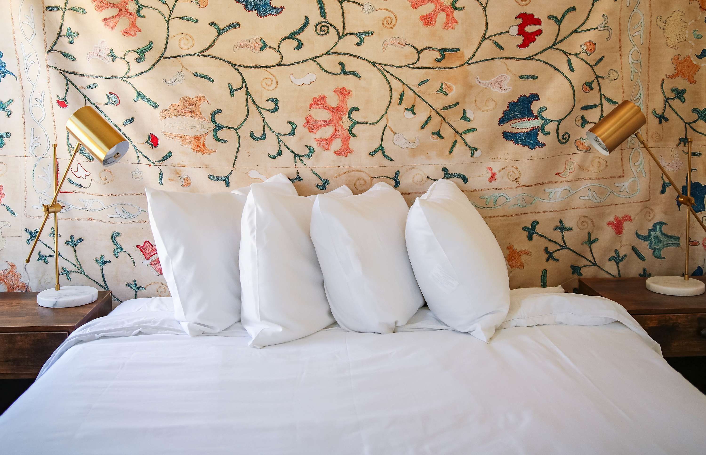 Een bed met witte beddengoed en meerdere kussens, een houten nachtkastje met een lamp, en een wand met bloementapijt.