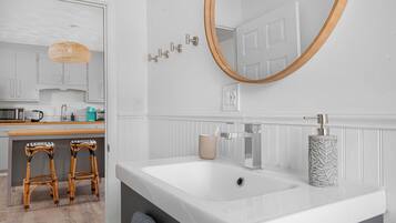 A modern bathroom with a white sink, a mirror with a wooden frame, and a toilet. There are two stools and a wicker lamp in the adjacent area.