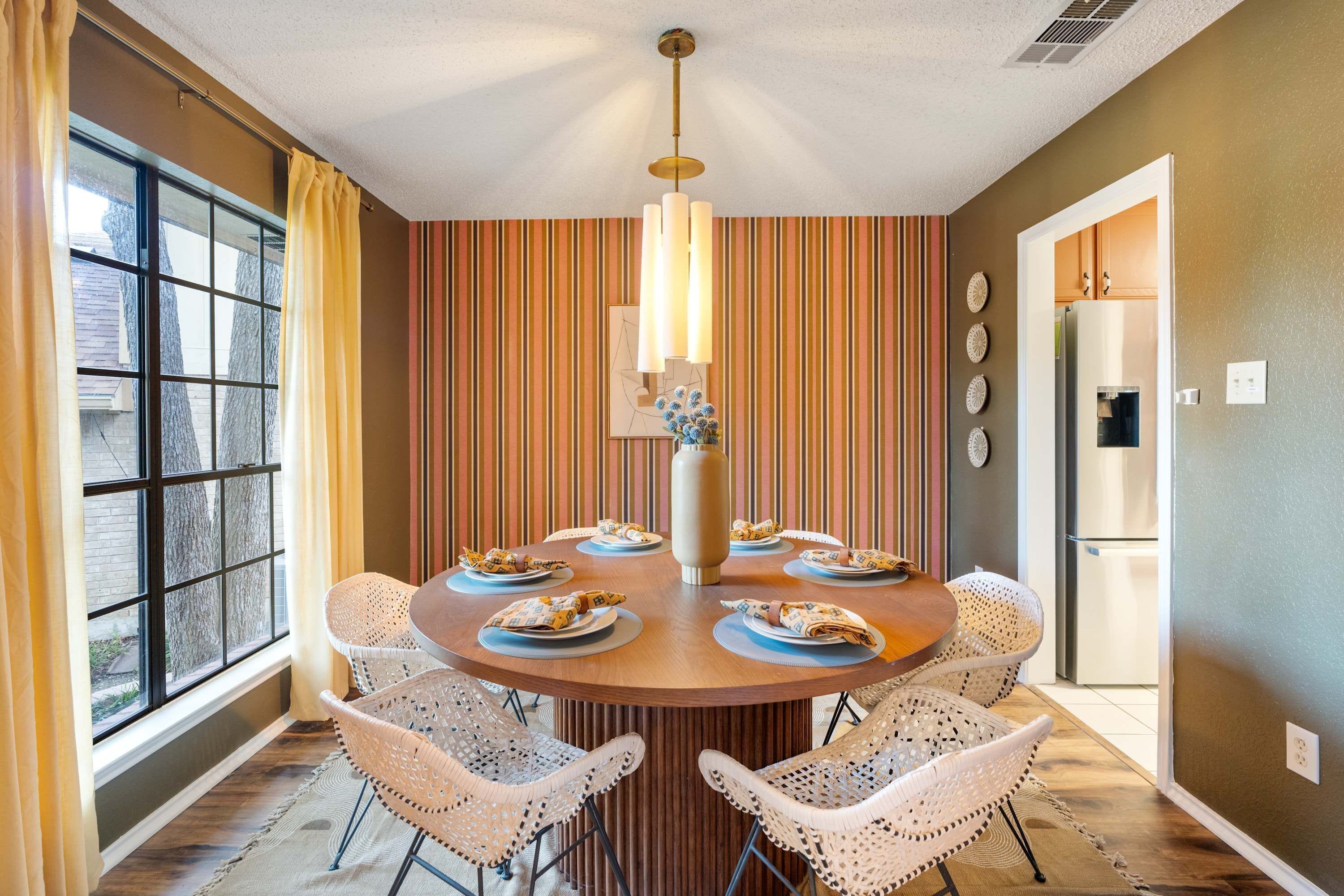 A dining area with a round table, six chairs, a vase with flowers, and a striped wall.