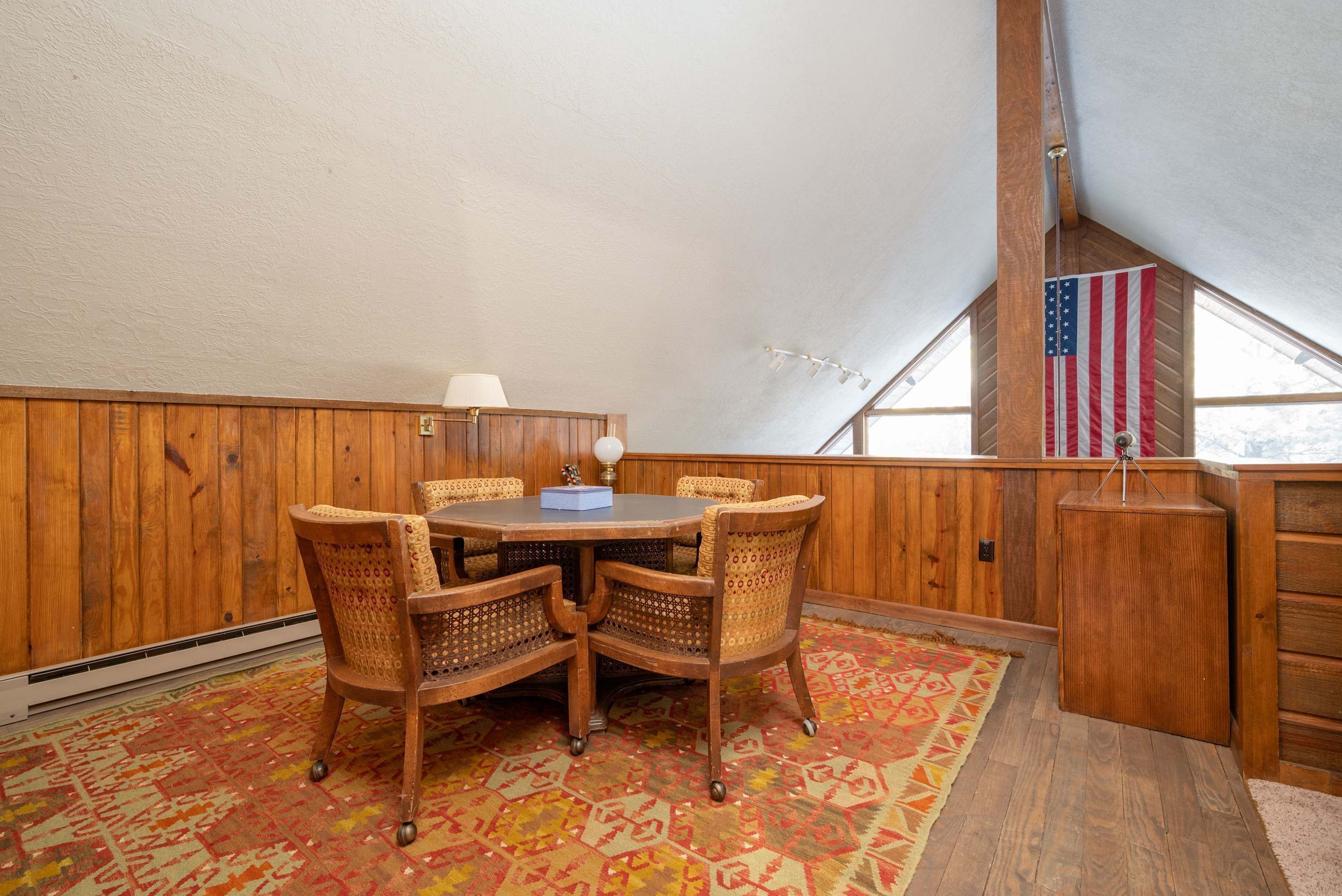 A room with a round wooden table, four wicker chairs, a patterned rug, a flag, and a slanted ceiling.