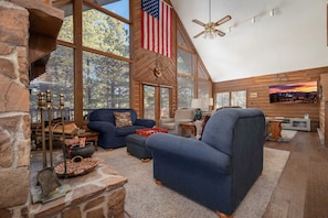 A spacious living room with a stone fireplace, large windows, and a high ceiling fan. - Pah s Haus Hot Tub Scenic View Dog-friendly (Angel Fire)