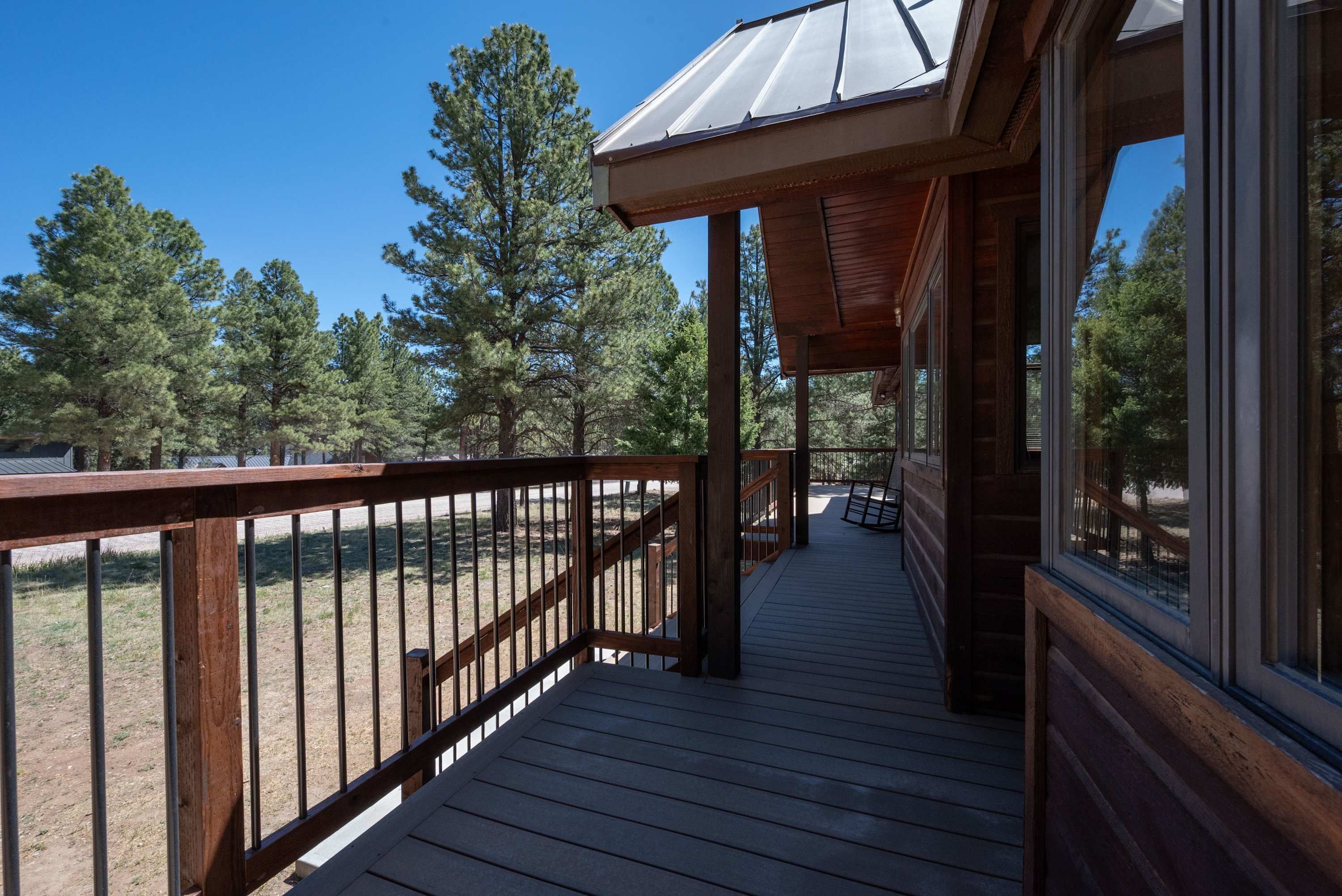 Une terrasse en bois avec une rambarde en métal, un espace couvert doté d’un toit en métal et une vue sur des pins en arrière-plan.