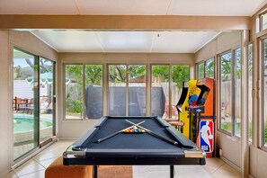 Ein Billardtisch mit Billardkielen und Kugeln, eine Nintendo 64 Spielkonsole und ein Basketballkorb in einem sonnendurchfluteten Raum mit großen Fenstern.