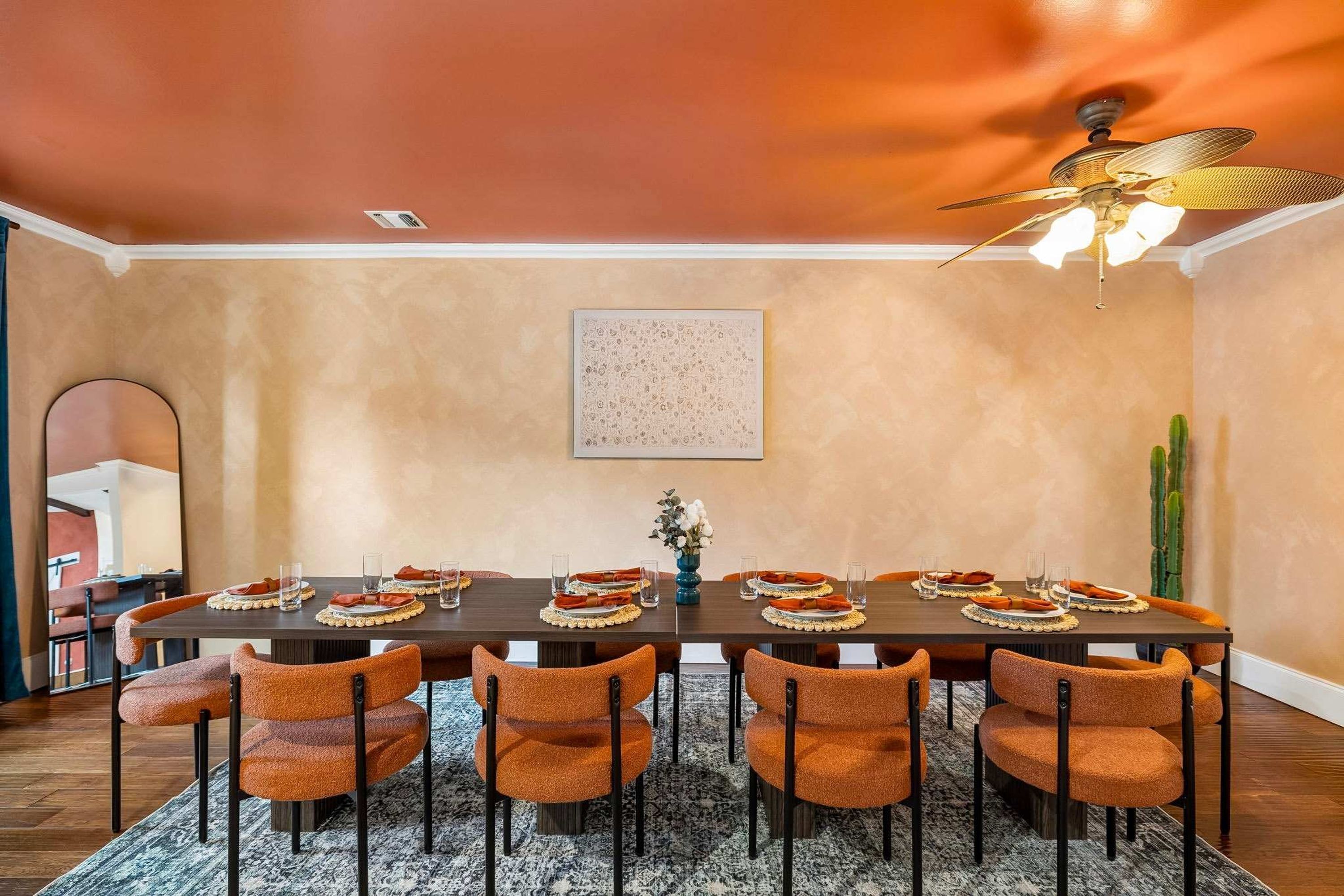 A dining room with a long table set for six, surrounded by six orange chairs, a ceiling fan, and a cactus plant.