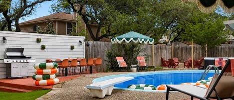 A backyard with a swimming pool, deck chairs, a barbecue grill, and a covered patio area.