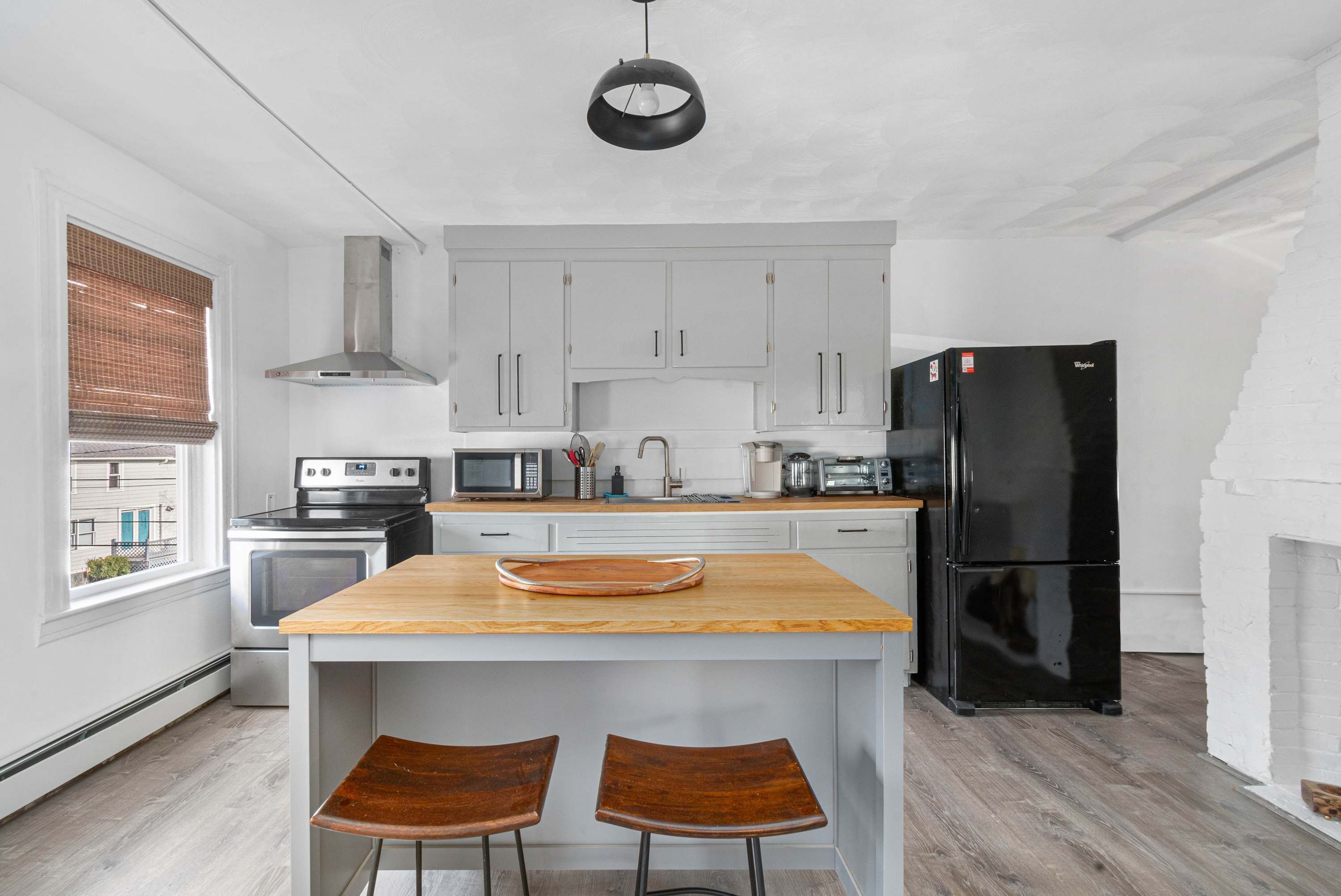 A modern kitchen with white cabinets, a black refrigerator, a wooden island with two stools, and a hanging light fixture.