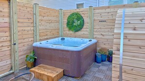 Un jacuzzi en un patio trasero con cerco de madera, cuenta con una terraza de madera, plantas en macetas y un banco de madera.