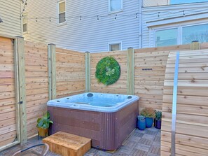 Un jacuzzi en un patio trasero con cerco de madera, cuenta con una terraza de madera, plantas en macetas y un banco de madera.