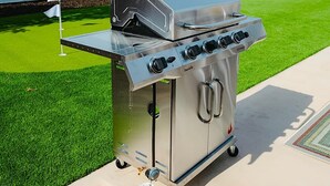 Ein tragbarer Grill aus Edelstahl mit integriertem Räucherofen und Seitenbrenner, auf einer Betonpatio mit Kunstgrasanlage platziert.