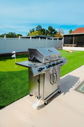 Ein tragbarer Grill aus Edelstahl mit integriertem Räucherofen und Seitenbrenner, auf einer Betonpatio mit Kunstgrasanlage platziert.