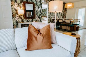 En hvid sofa med en strukturfarvet pude, et spejl, en lampe og et spiseområde med stole og et bord.
