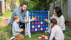 En familj som spelar ett brädspel utomhus, med ett träbord och en gräsplätt i bakgrunden.