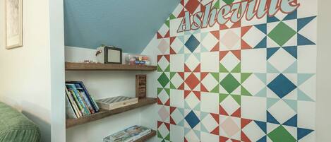 Una habitación con un papel tapiz de diseño geométrico, estantes con libros y juguetes, y un cuadro en la pared.