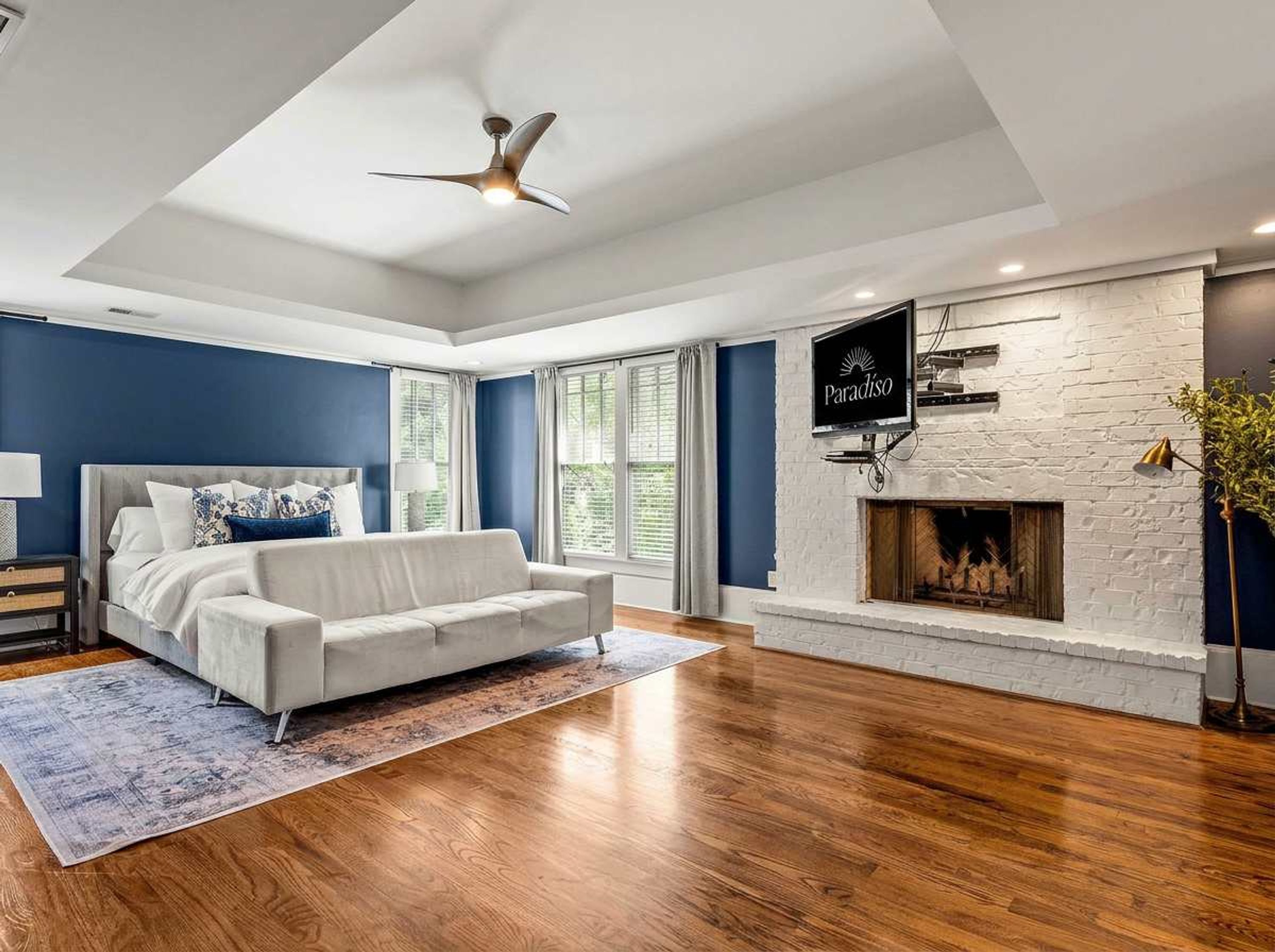 A modern bedroom with a bed, a white sofa, a fireplace, and a ceiling fan.