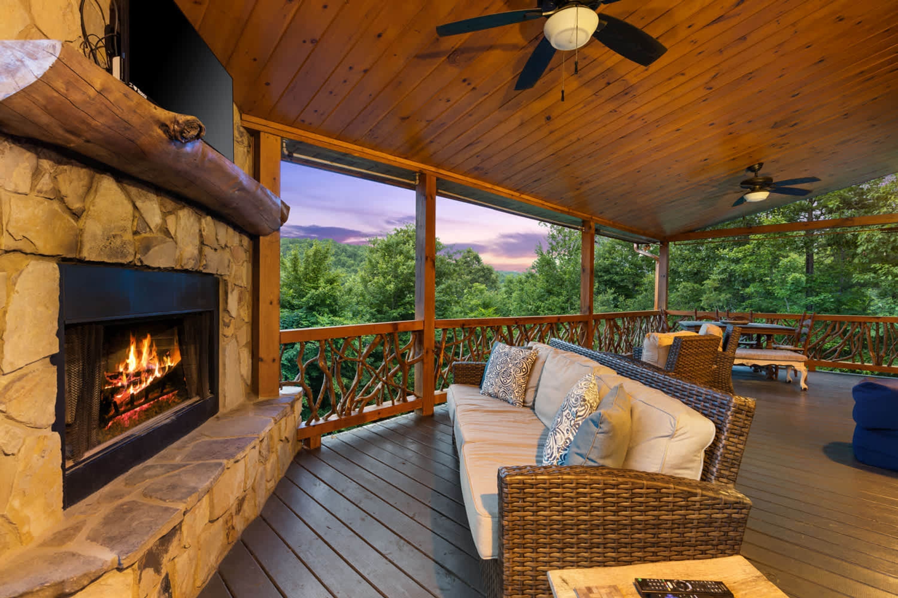 A covered outdoor area with a stone fireplace, wicker furniture, and a view of the forest.