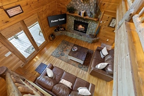 Interior acolhedor de uma cabana com lareira de pedra, sofás de couro e uma televisão de tela plana com a inscrição 'Home Team'.