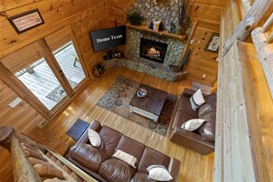 Interior acogedor de una cabaña con chimenea de piedra, sofás de cuero y un televisor de pantalla plana con la etiqueta 'Home Team'.