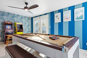 A pool table with a ceiling fan, arcade games, and framed pictures on a blue wall.