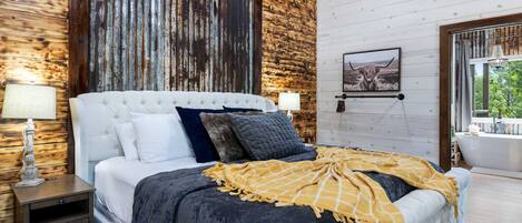 A bedroom with a large bed, a nightstand, a lamp, a wooden wall, a framed picture, and a window with a view.